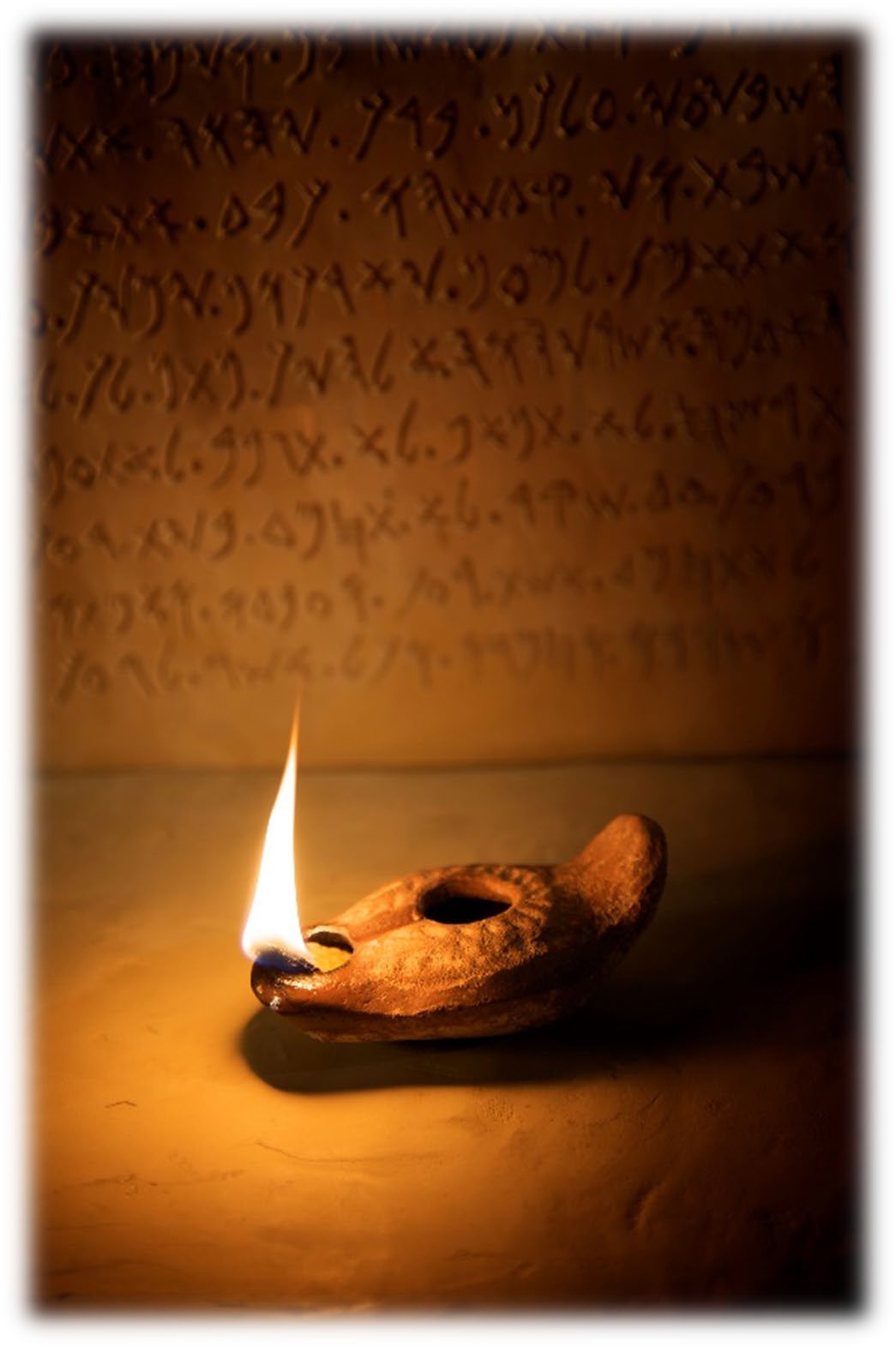 A small clay oil lamp burning with a single flame on a stone surface, with lines of ancient Hebrew script on the wall behind it
