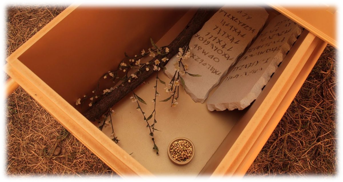 Looking down into an open gold-lined chest holding two inscribed stone tablets, a blossoming almond branch and a small dish of manna