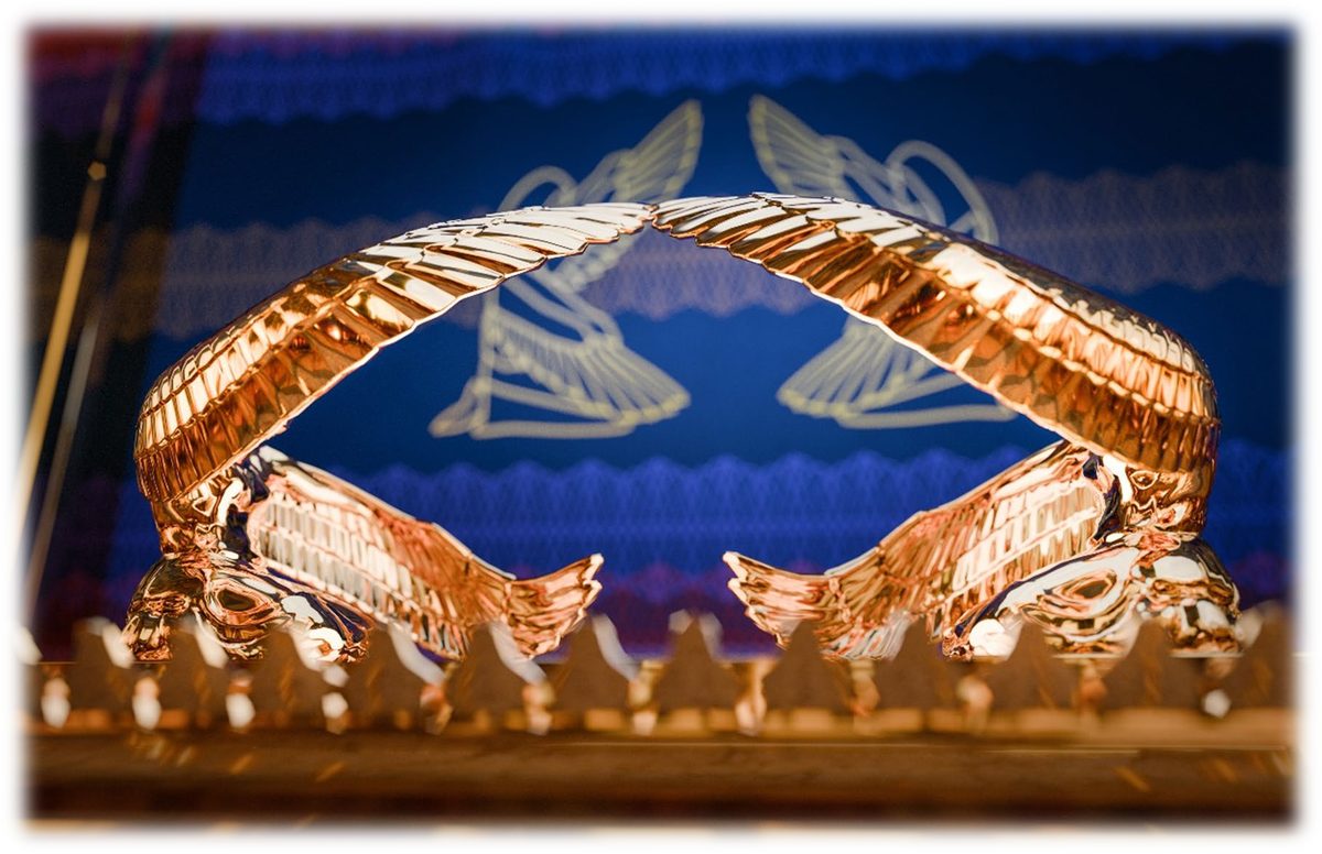 Two golden cherubim on the ends of the Mercy Seat, their wings lifted up and meeting over it, with a blue embroidered curtain behind